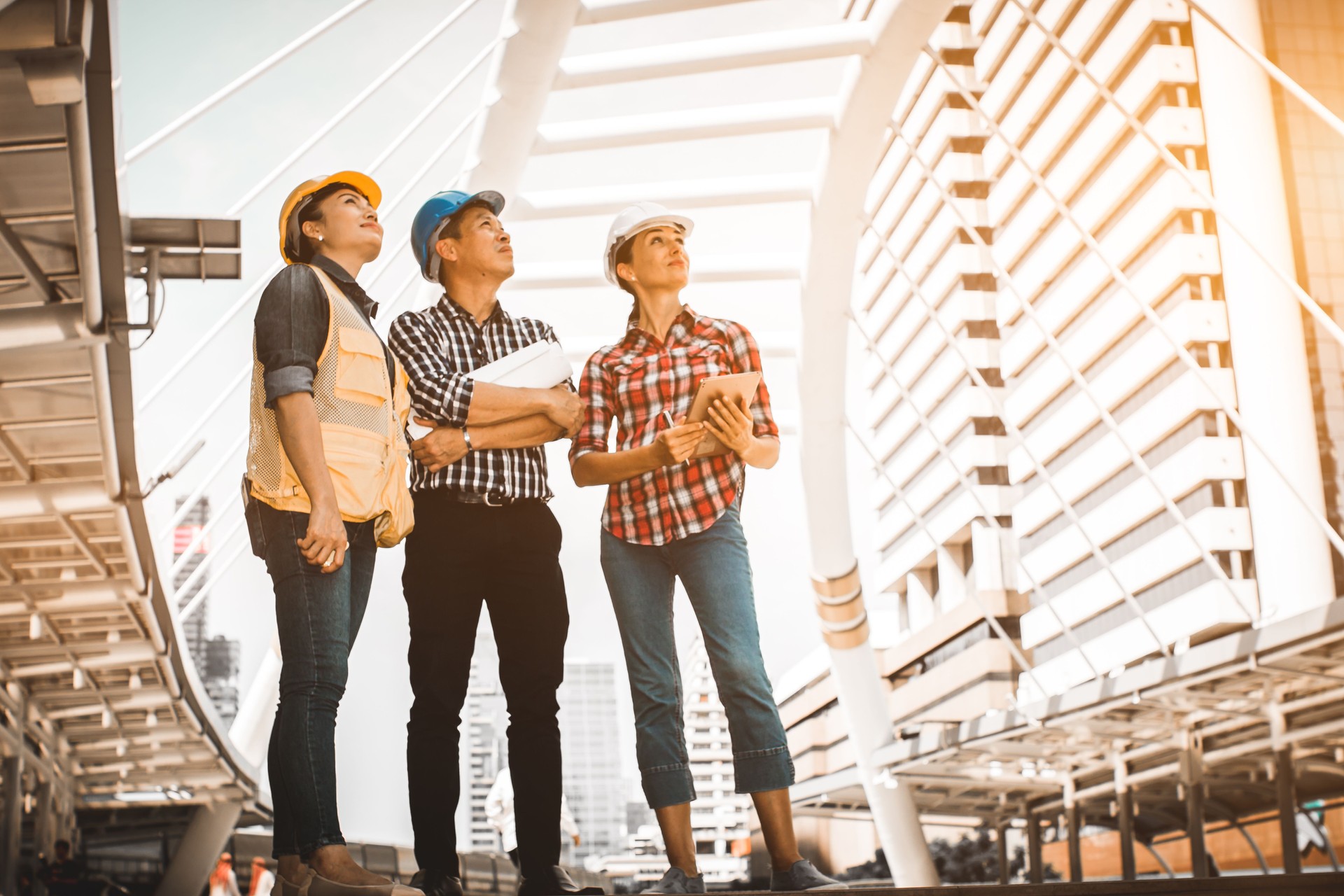 Three industrial engineer standing and looking to right side wear safety helmet with holding inspection and digital tablet on building outside. Engineering tools and construction concept.